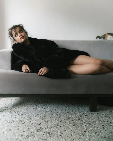 Young woman in a black fur coat rests on a gray couch in an indoor setting, accentuated by cool, natural light and a terrazzo floor.