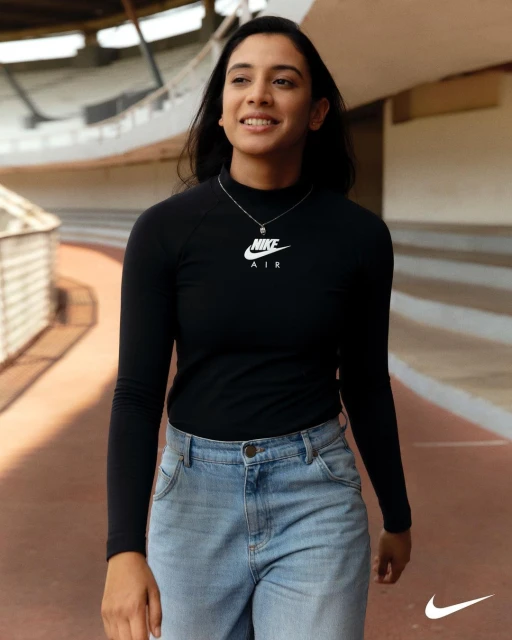 A young adult woman in a long-sleeved shirt and jeans walks along an outdoor track. She wears a Nike shirt and pendant necklace.