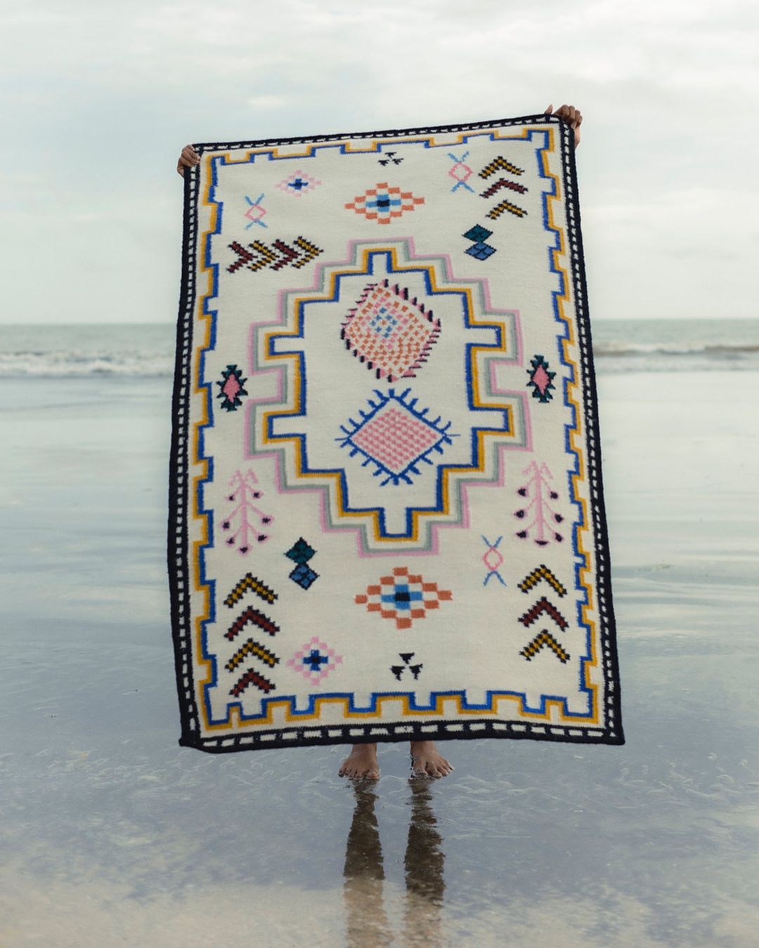 Person holding patterned rug at beach with water reflections. Rug displays tribal style geometric shapes and soft color palette.