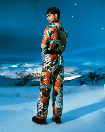 Young man poses in a patterned ski suit against a snowy, mountain backdrop under a blue and starry sky, captured in a fashion shot.