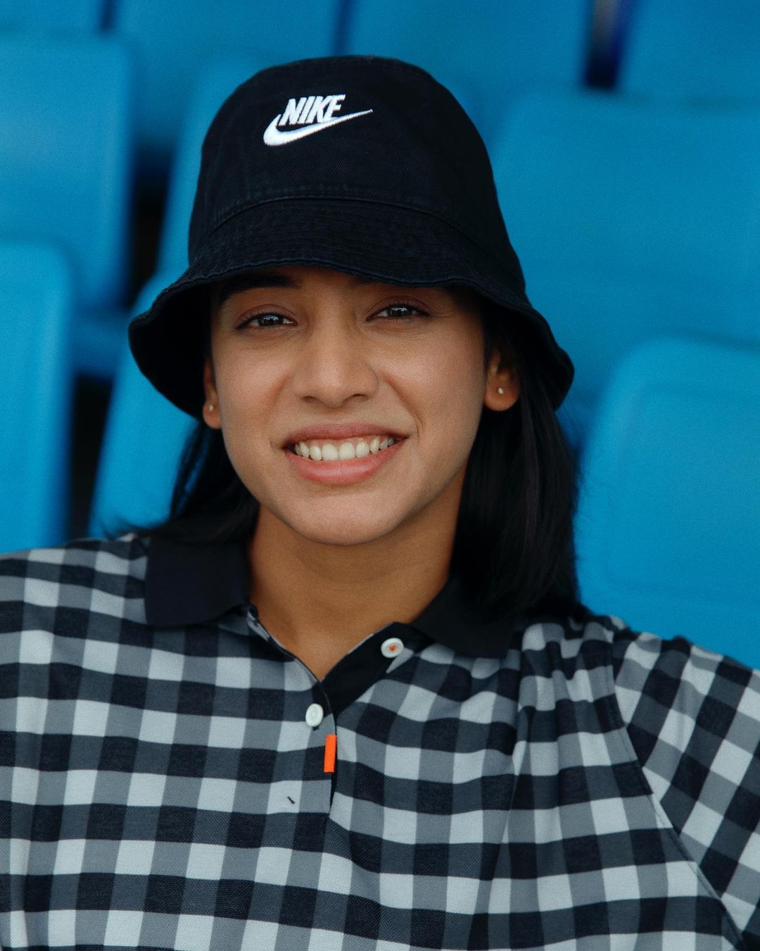 Smiling adult female wearing a black and white patterned polo shirt and Nike bucket hat sits in front of blue stadium seats.