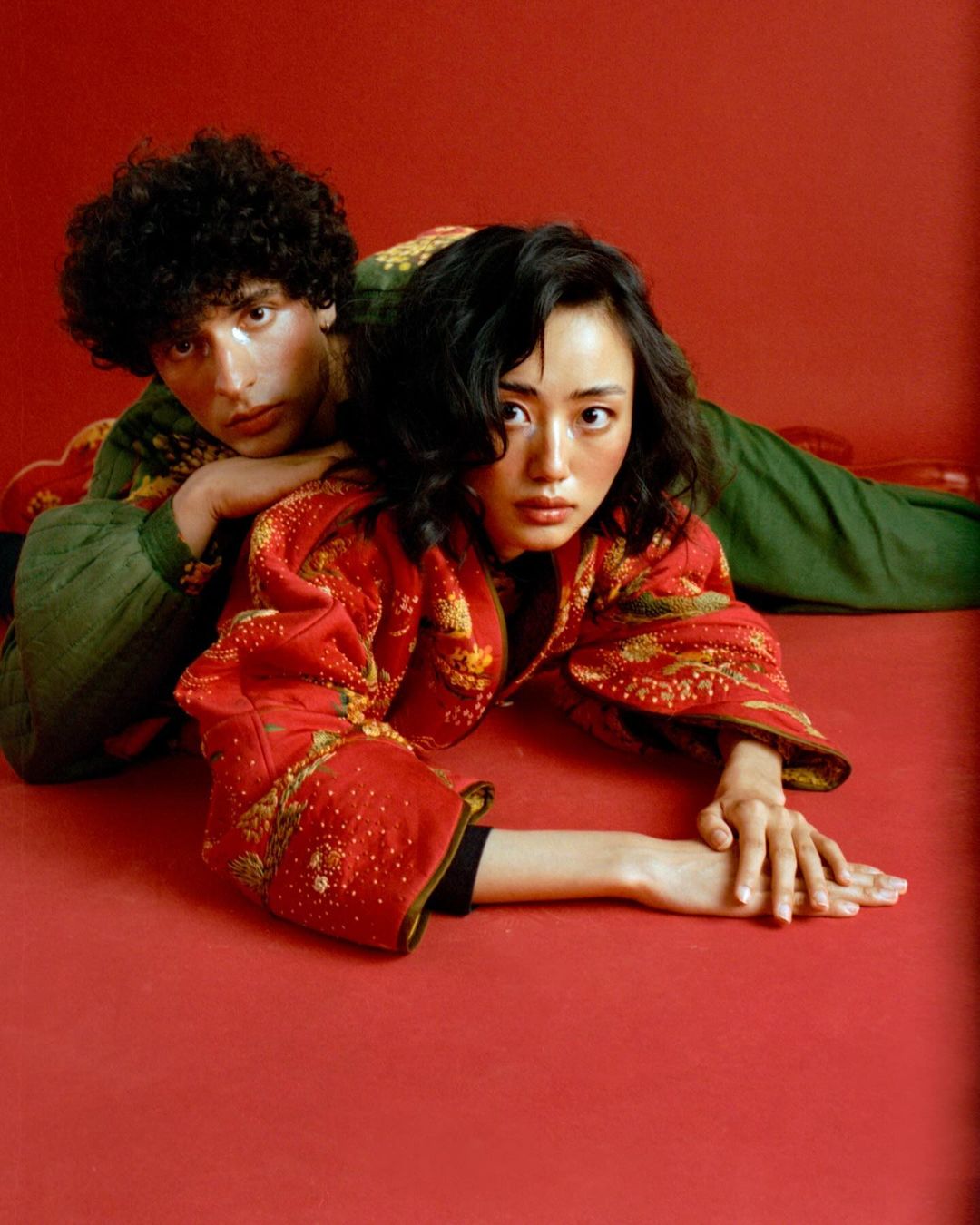 Asian female model in red patterned jacket poses with a curly haired man in green patterned jacket laying on red seamless background.