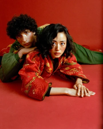 Asian female model in red patterned jacket poses with a curly haired man in green patterned jacket laying on red seamless background.