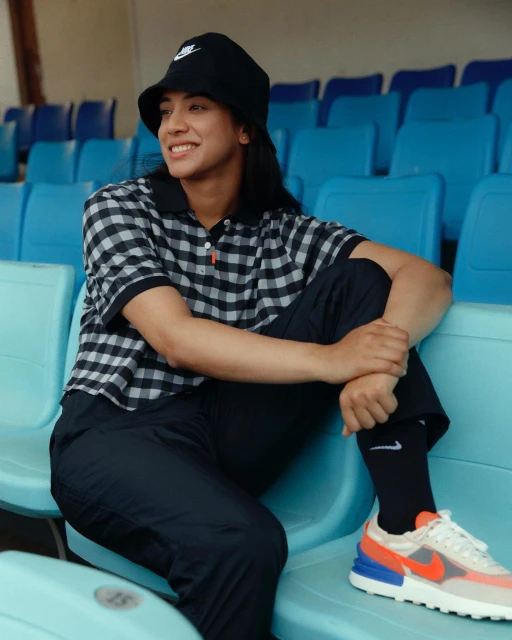 A woman in a bucket hat and checkered shirt sits on blue stadium seats, wearing Nike shoes and socks, smiling in natural light.