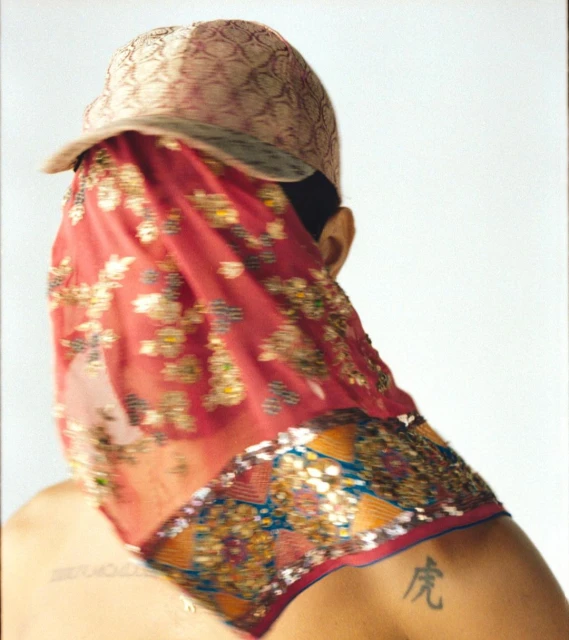 Person wearing decorated baseball cap and sequined veil against white backdrop, showcasing fashion and cultural fusion.