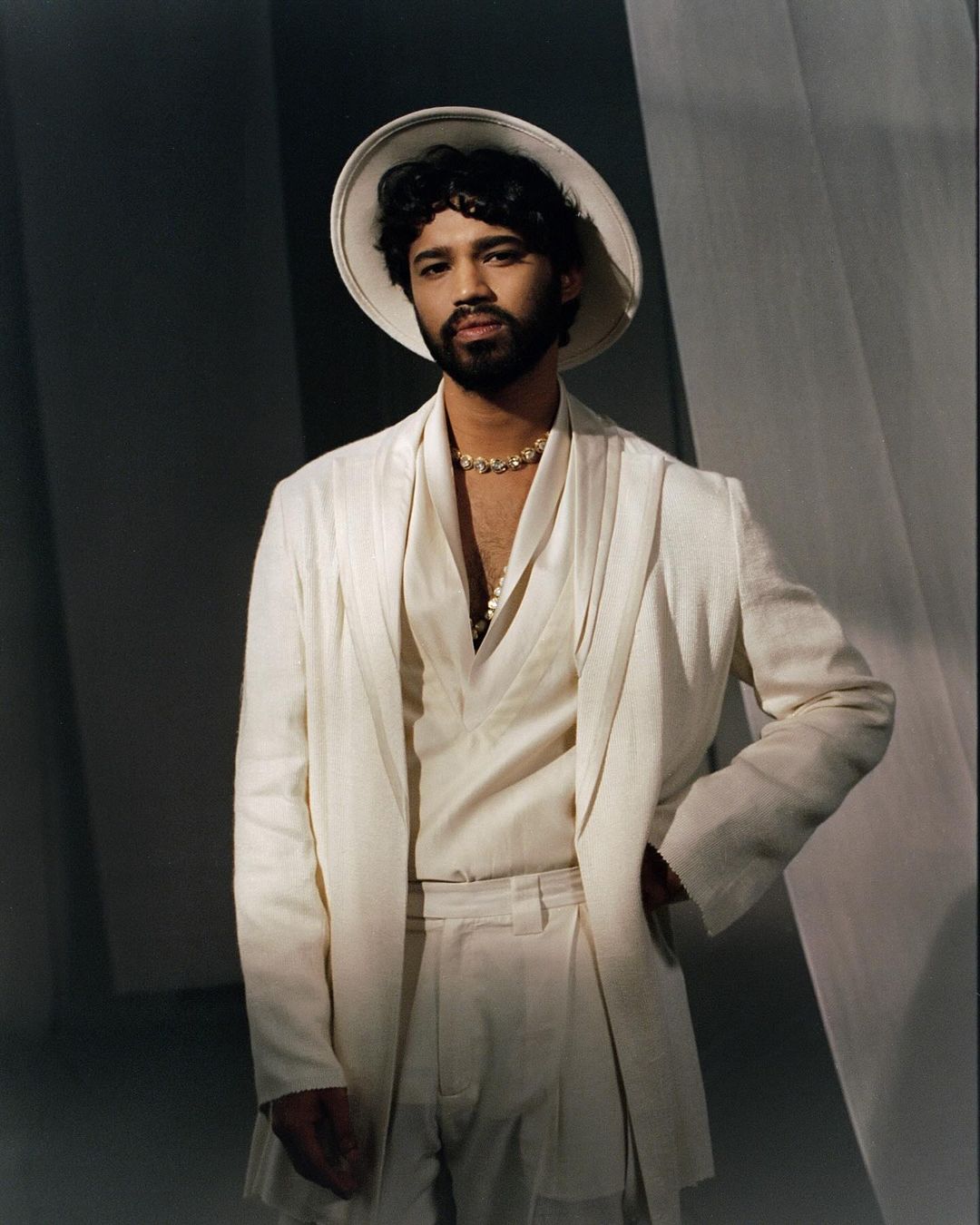 Man poses in an all-white suit with a hat against a light backdrop, exuding a sophisticated and stylish aesthetic in a studio setting.