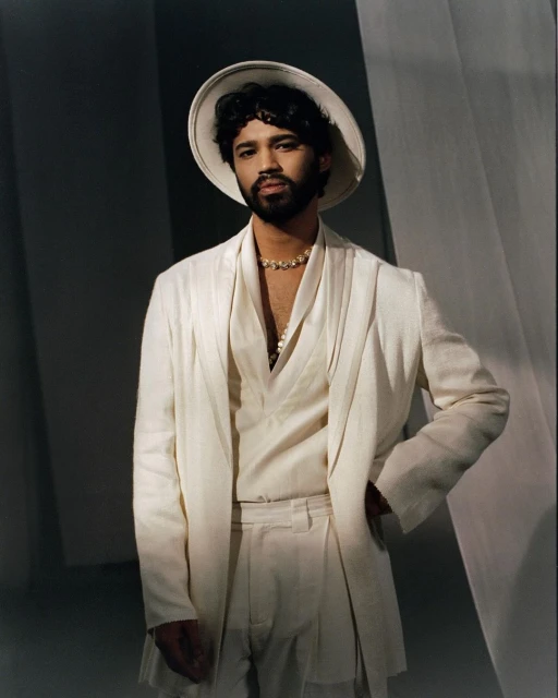 Man poses in an all-white suit with a hat against a light backdrop, exuding a sophisticated and stylish aesthetic in a studio setting.