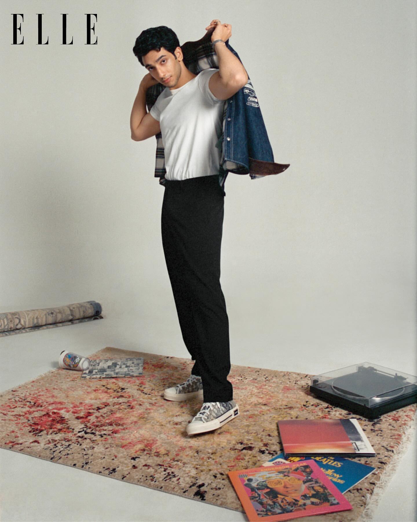 Portrait of a young adult male posing in a studio with a jacket over his shoulder, wearing a white t-shirt and black trousers, with albums on the floor.