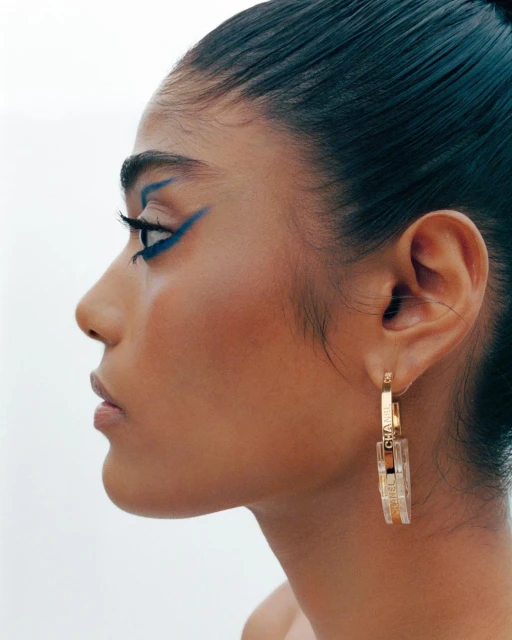 Close-up profile of a woman with blue eyeliner and gold Chanel hoop earrings against a bright white background.