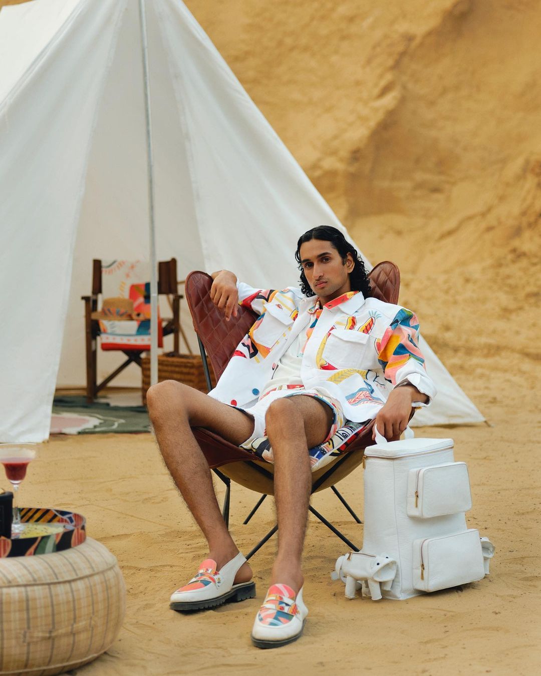 A dark-haired man in a patterned jacket and shorts sits in a chair outside of a tent on a sandy surface with a cooler next to him.