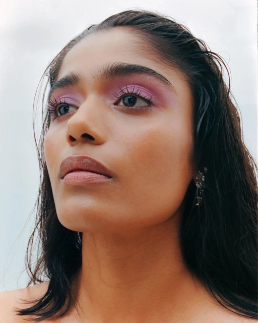 Close-up of a young woman with lilac eyeshadow and dark hair against a light background, looking off camera to the left side.