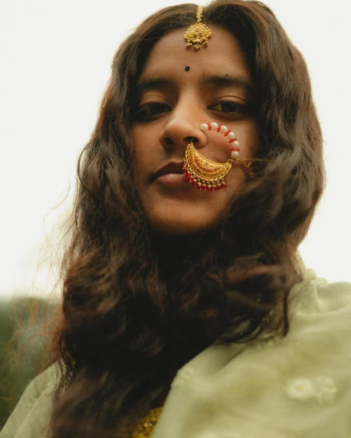 Portrait of a South Asian woman with traditional gold jewelry, including a nose ring and maang tikka, against a bright outdoor backdrop.