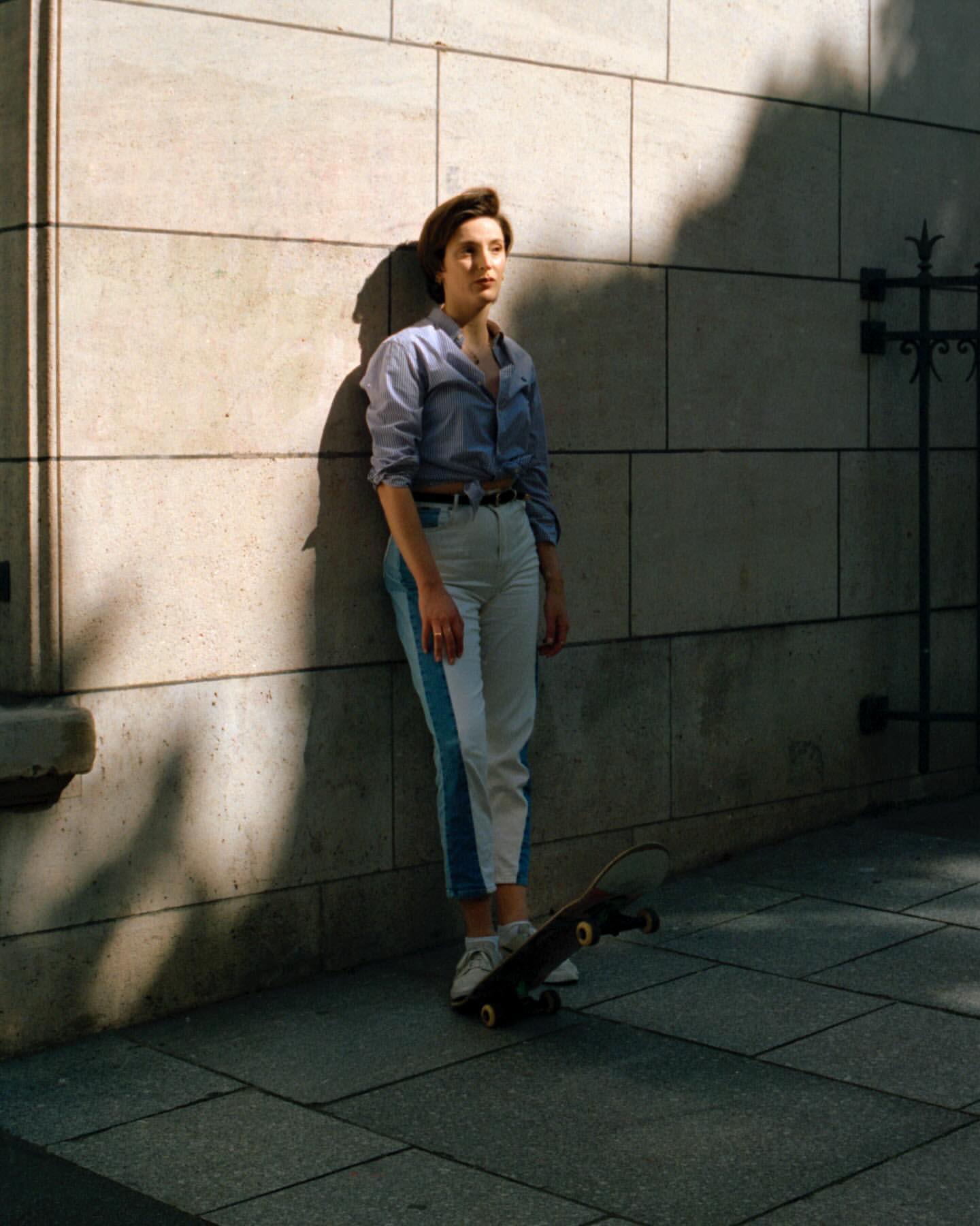 Woman with short hair leans against a stone wall, skateboard at her feet. Sunlight and shadow play across the textured surface.