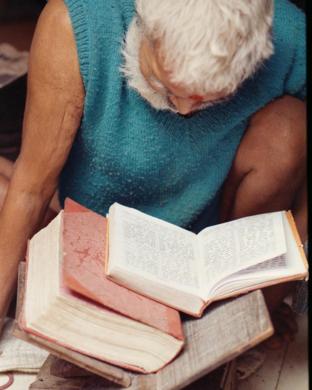 Elderly man with white hair and blue vest sitting while reading a book. Books are stacked on a wooden surface.