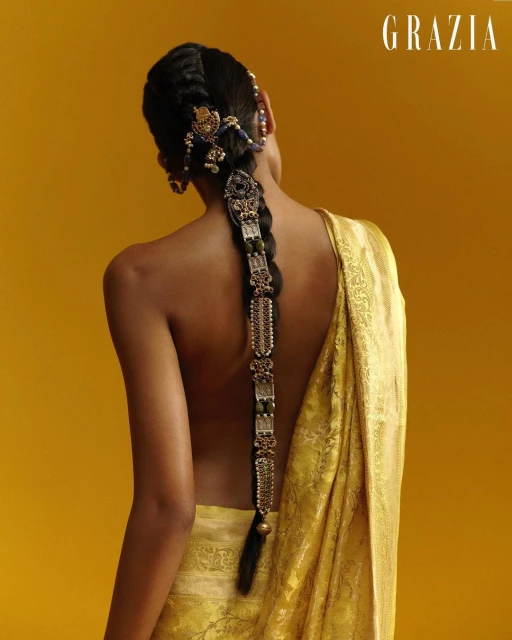 Elegant back view of a woman with a long, ornate braid adorned with jewels, wearing a golden patterned shawl against a yellow background.