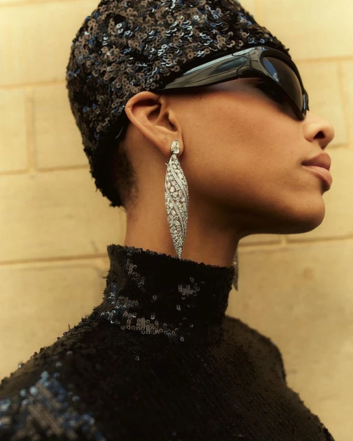 Elegant woman with dark complexion poses wearing sequined top, hat, statement earrings and sunglasses in a sophisticated indoor setting.