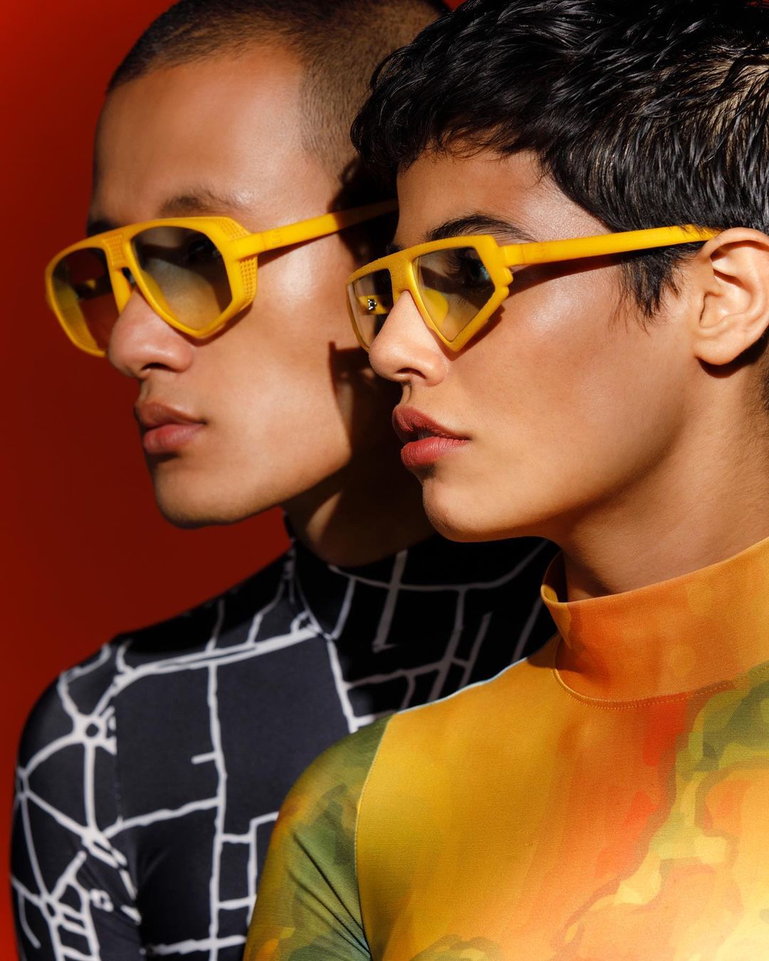 Two people wearing yellow geometric sunglasses and patterned turtlenecks pose against a red backdrop in a studio setting, fashion portrait.