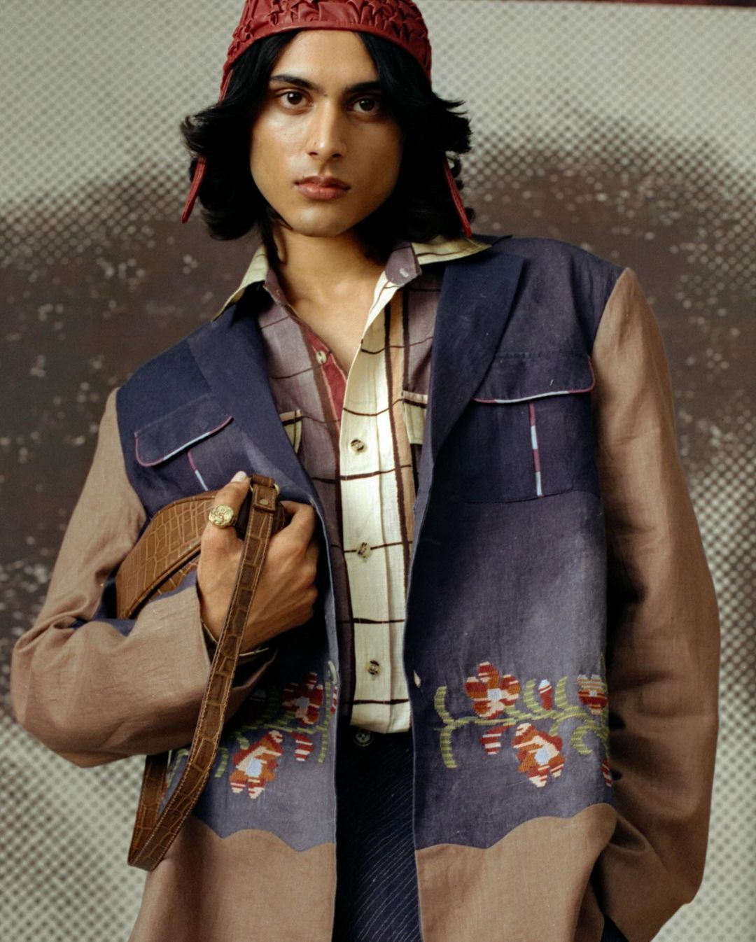 Male model poses in a patterned brown and blue blazer, collared shirt, knit hat, and dark pants, holding a brown leather purse indoors.