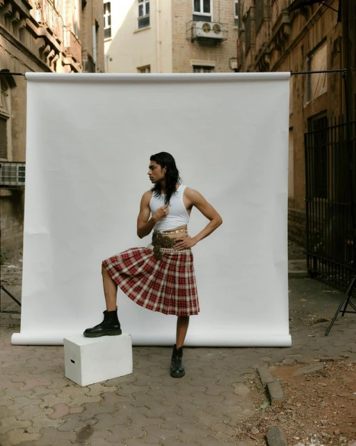 Young man poses in a white crop top, plaid kilt, and black boots, standing on a white block against a white backdrop in an alley.