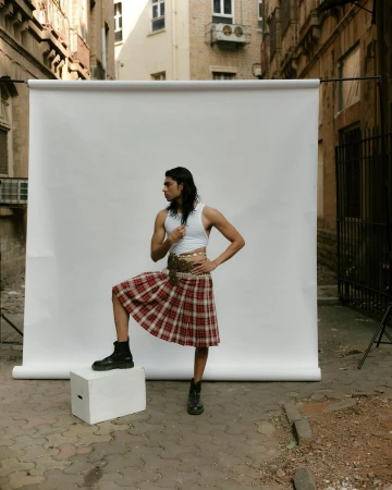 Young man poses in a white crop top, plaid kilt, and black boots, standing on a white block against a white backdrop in an alley.