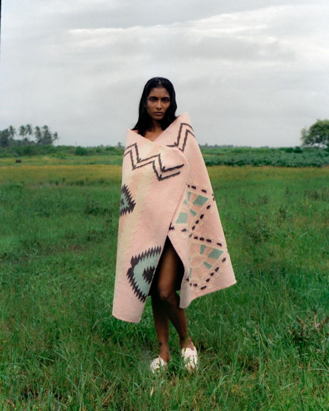 A woman wrapped in a patterned blanket stands in a grassy field under an overcast sky, wearing white shoes.