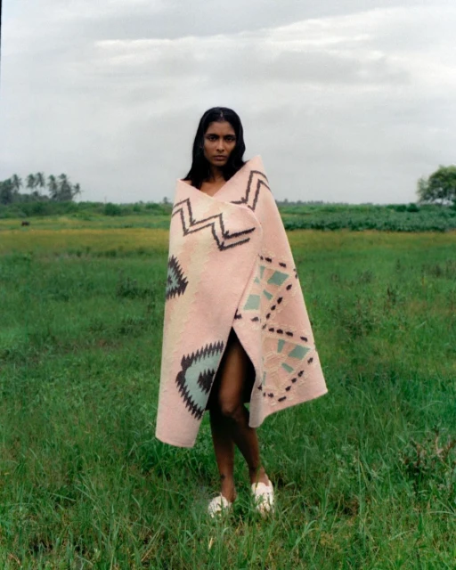 A woman wrapped in a patterned blanket stands in a grassy field under an overcast sky, wearing white shoes.
