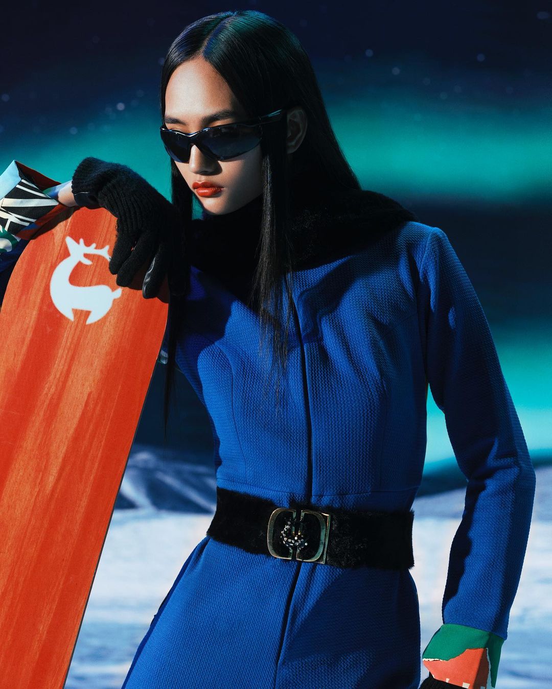 An Asian woman in a blue jumpsuit and sunglasses poses in a studio with a snowboard featuring a reindeer logo.