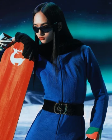 An Asian woman in a blue jumpsuit and sunglasses poses in a studio with a snowboard featuring a reindeer logo.