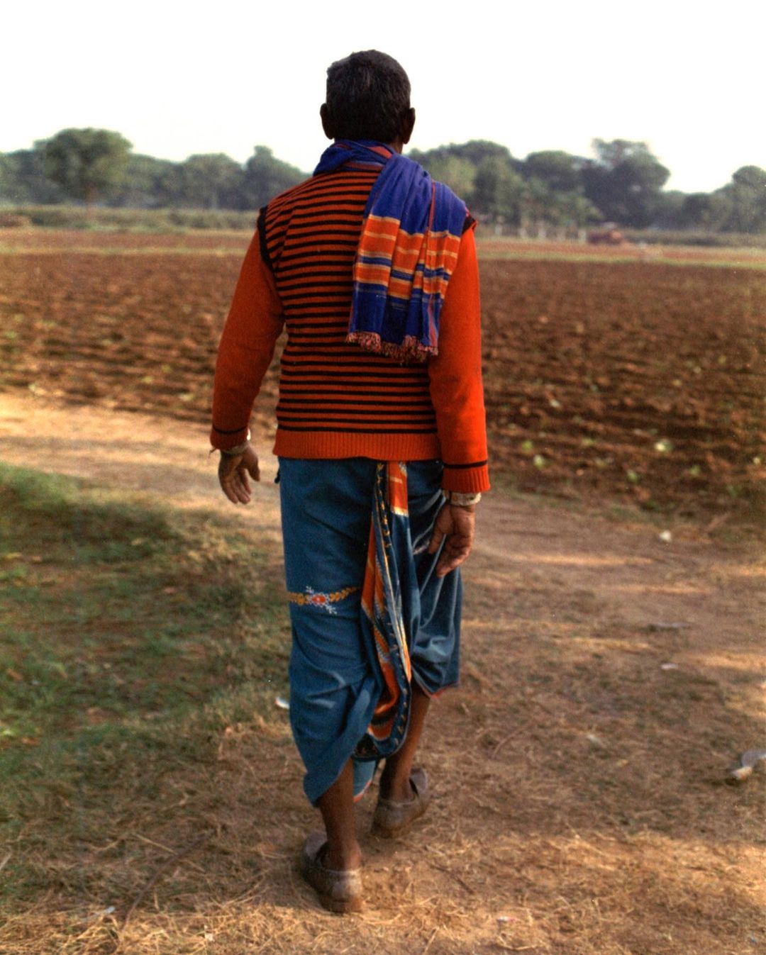 An adult male walks through a rural field in traditional clothing, showcasing a back view with trees and sky in the background.