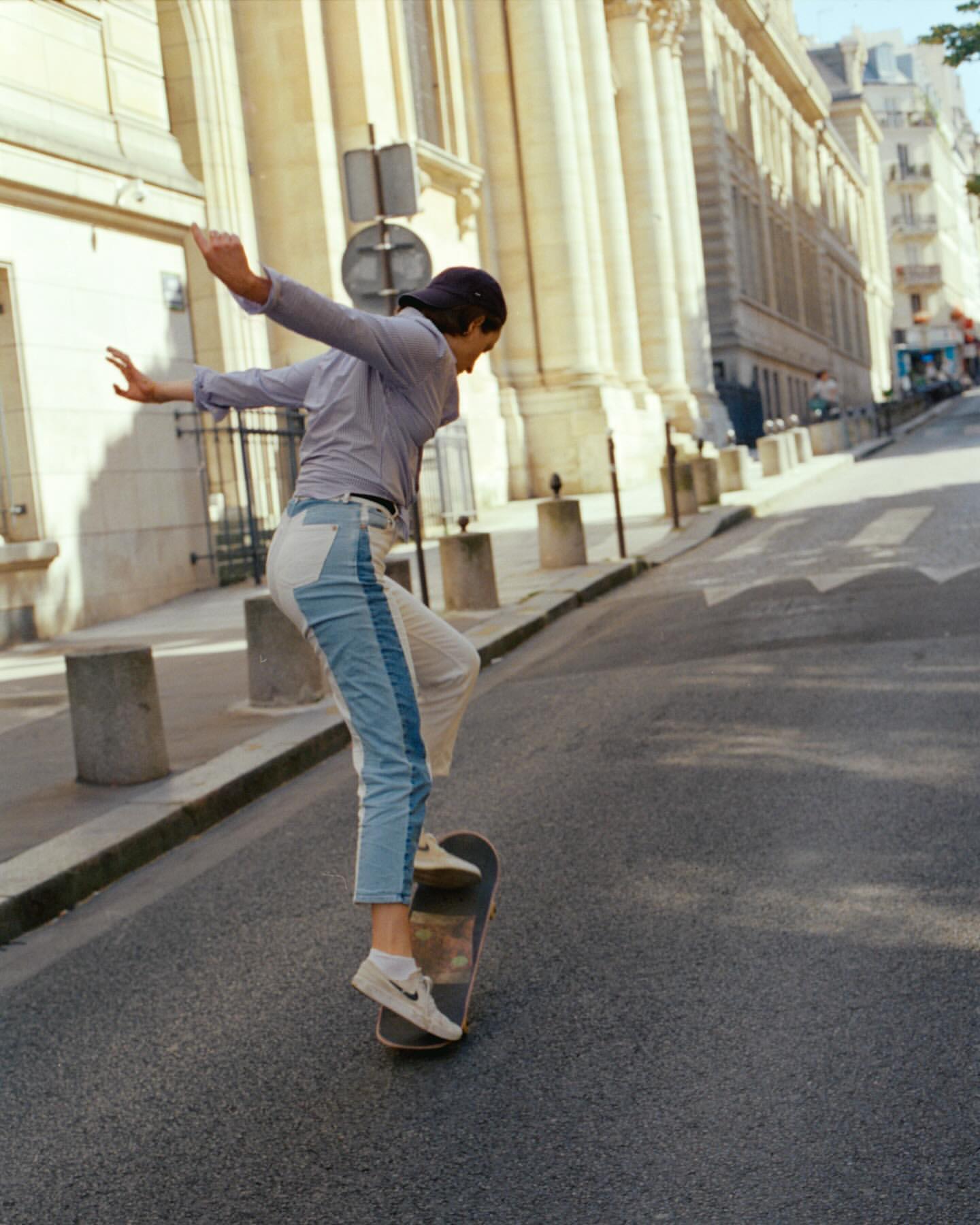 Young adult woman skateboarding down a city street in daytime. She wears jeans and a long sleeve shirt with buildings in the background.