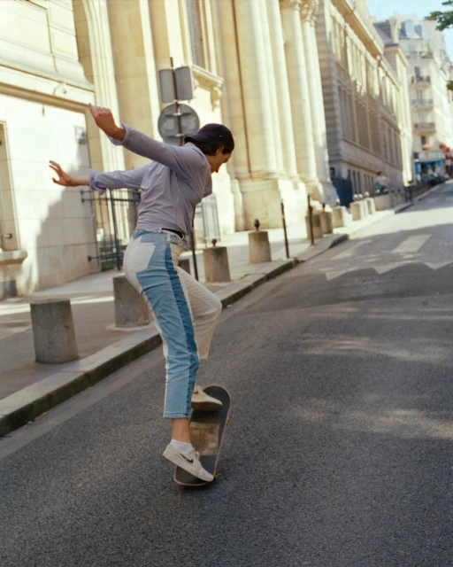 Young adult woman skateboarding down a city street in daytime. She wears jeans and a long sleeve shirt with buildings in the background.