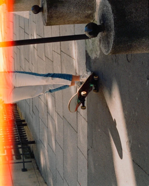 A skateboard rests on a paved surface alongside a person's legs, featuring concrete architectural elements and a metal bench.