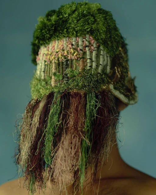 Person wearing a handmade knitted hat with tree design and yarn strands against a blue background in a studio setting.