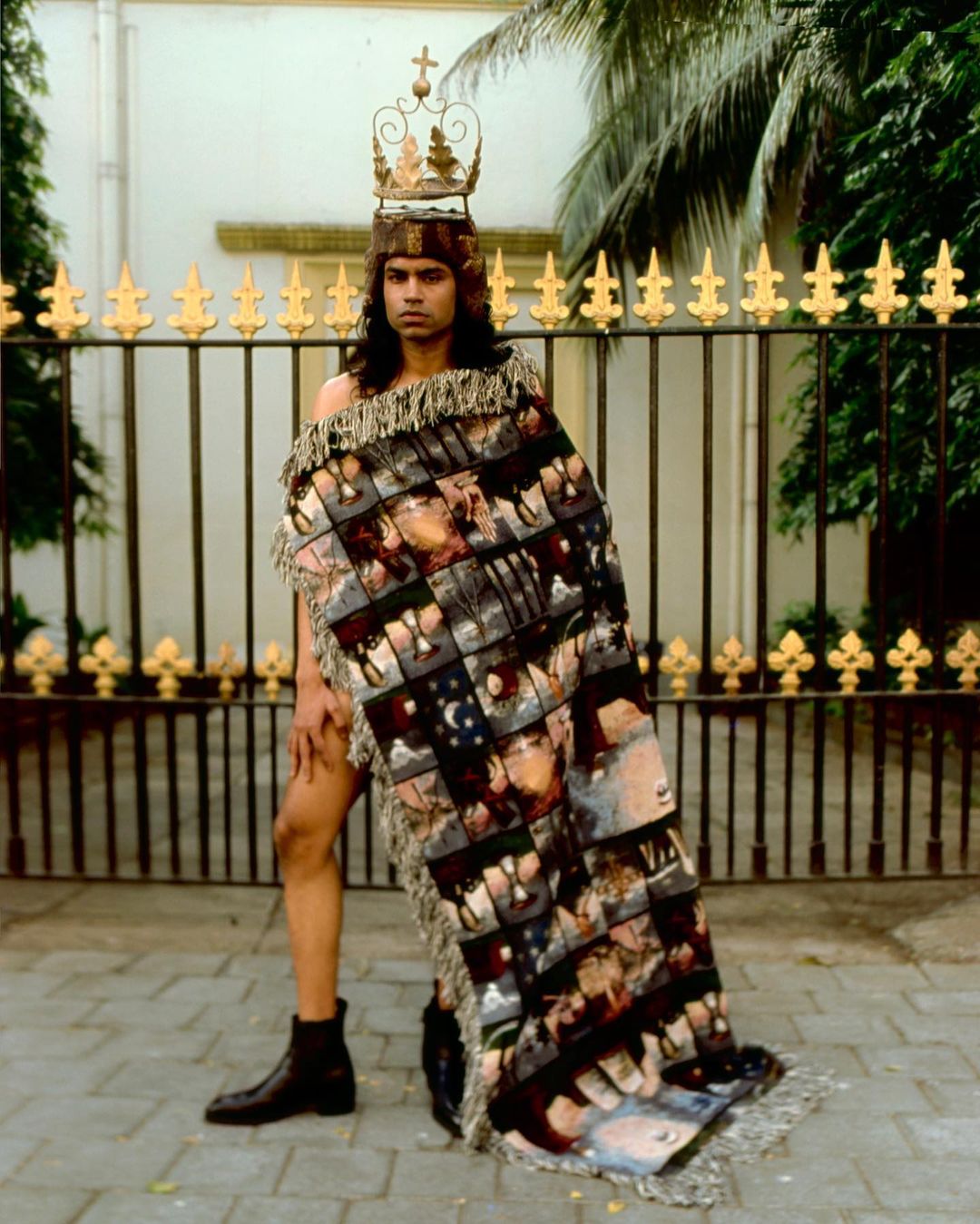 Man wears decorative headpiece and draped patterned fabric posing in front of gate and building in outdoor setting.