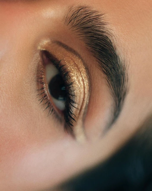 Close-up studio shot of an adult female's eye with gold-colored eyeshadow, dark lashes, and a neatly groomed eyebrow.