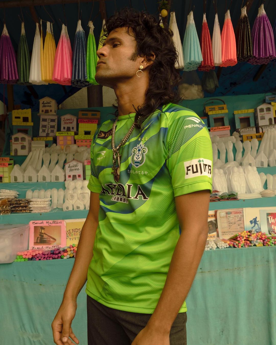 Adult male wearing a green soccer jersey standing in front of colorful toy houses and lamps in an indoor marketplace setting.