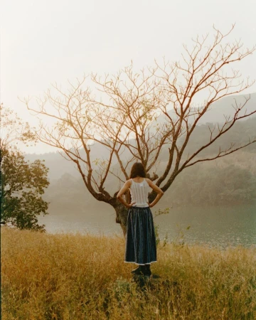 Rear view of a person standing with hands on hips before a bare tree in a dry grassy field overlooking a calm body of water.