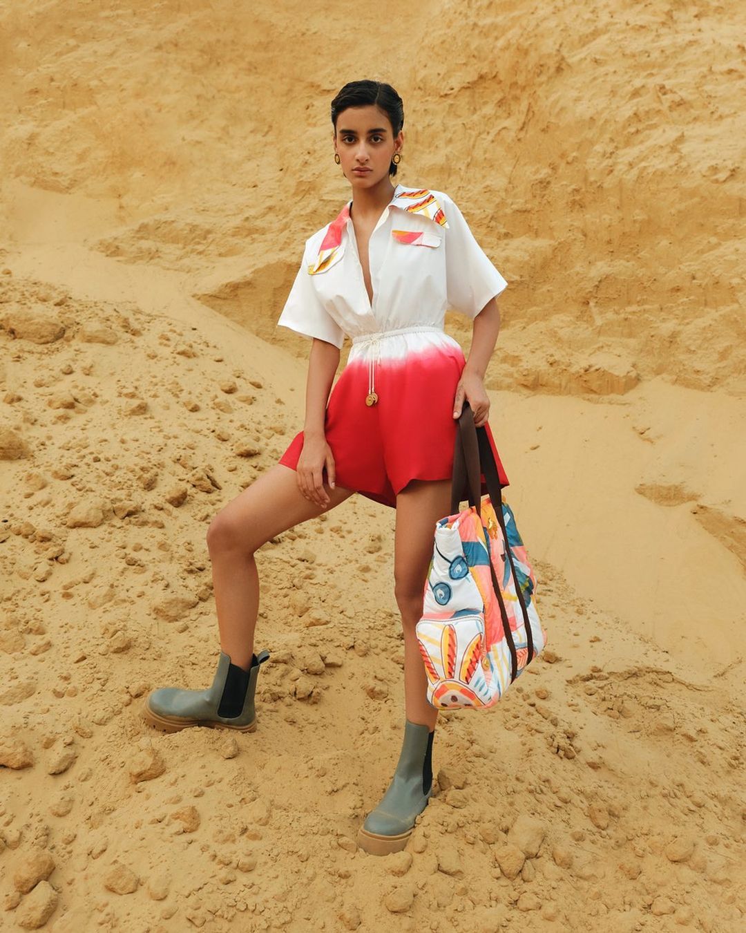 Woman poses in a white short-sleeve collared shirt with orange accents and red shorts in a sandy, outdoor environment with grey boots.
