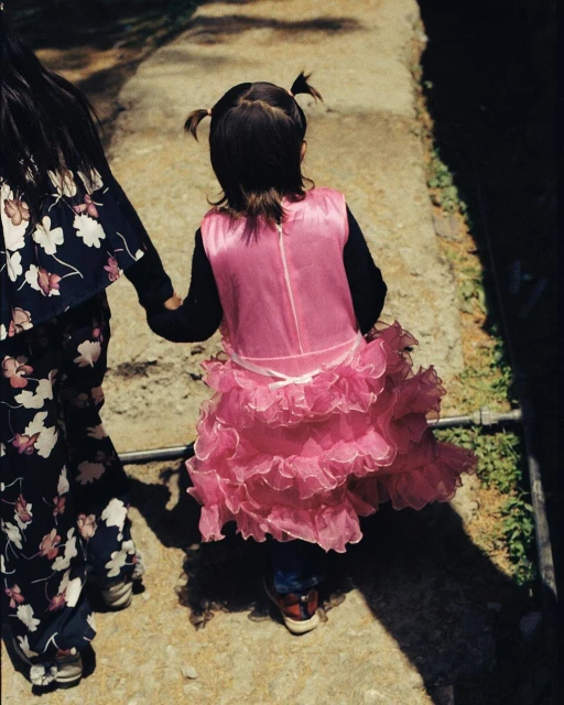 A young child in a pink dress holds hands with an adult on a sunny path, viewed from behind, showcasing a sweet moment outdoors.