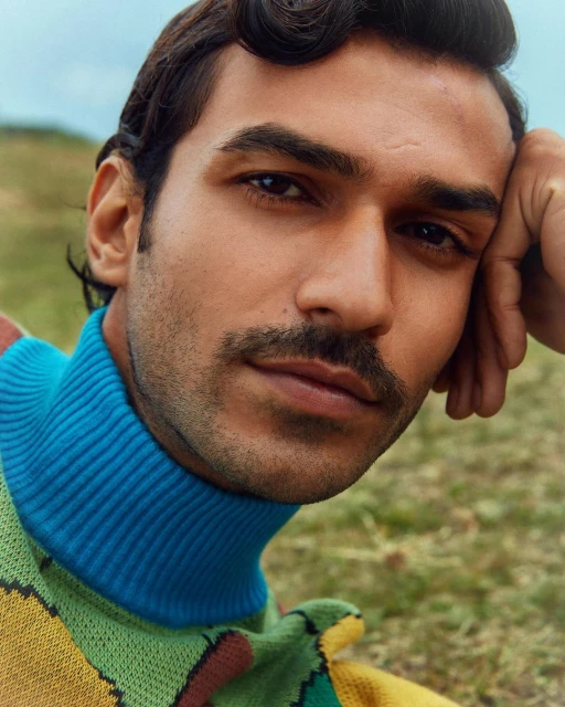 Close-up portrait of a dark-haired adult male with a moustache, wearing a multi-colored sweater in an outdoor environment with green grass.