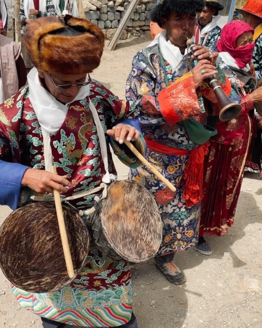 Two musicians in traditional Tibetan clothing play instruments outdoors; one plays a drum, the other plays a wind instrument at an event.
