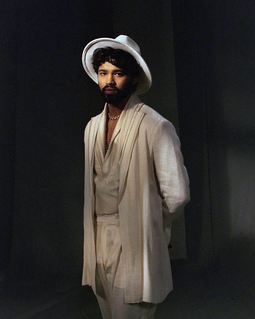 Portrait of an adult male wearing a white suit and a wide-brimmed hat, standing indoors with a neutral facial expression, against a dark background.
