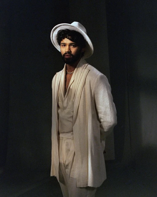 Portrait of an adult male wearing a white suit and a wide-brimmed hat, standing indoors with a neutral facial expression, against a dark background.