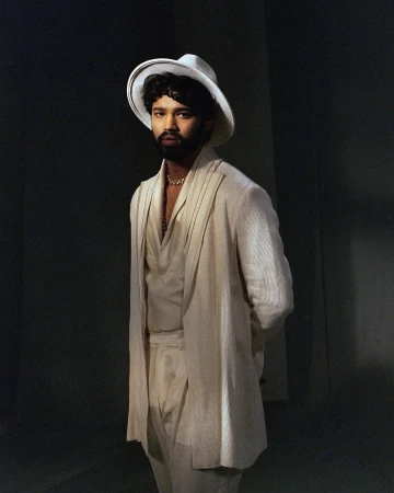 Portrait of an adult male wearing a white suit and a wide-brimmed hat, standing indoors with a neutral facial expression, against a dark background.