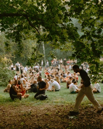 A diverse group of people gathers outdoors on a grassy field under trees, with some sitting and others walking in daylight.