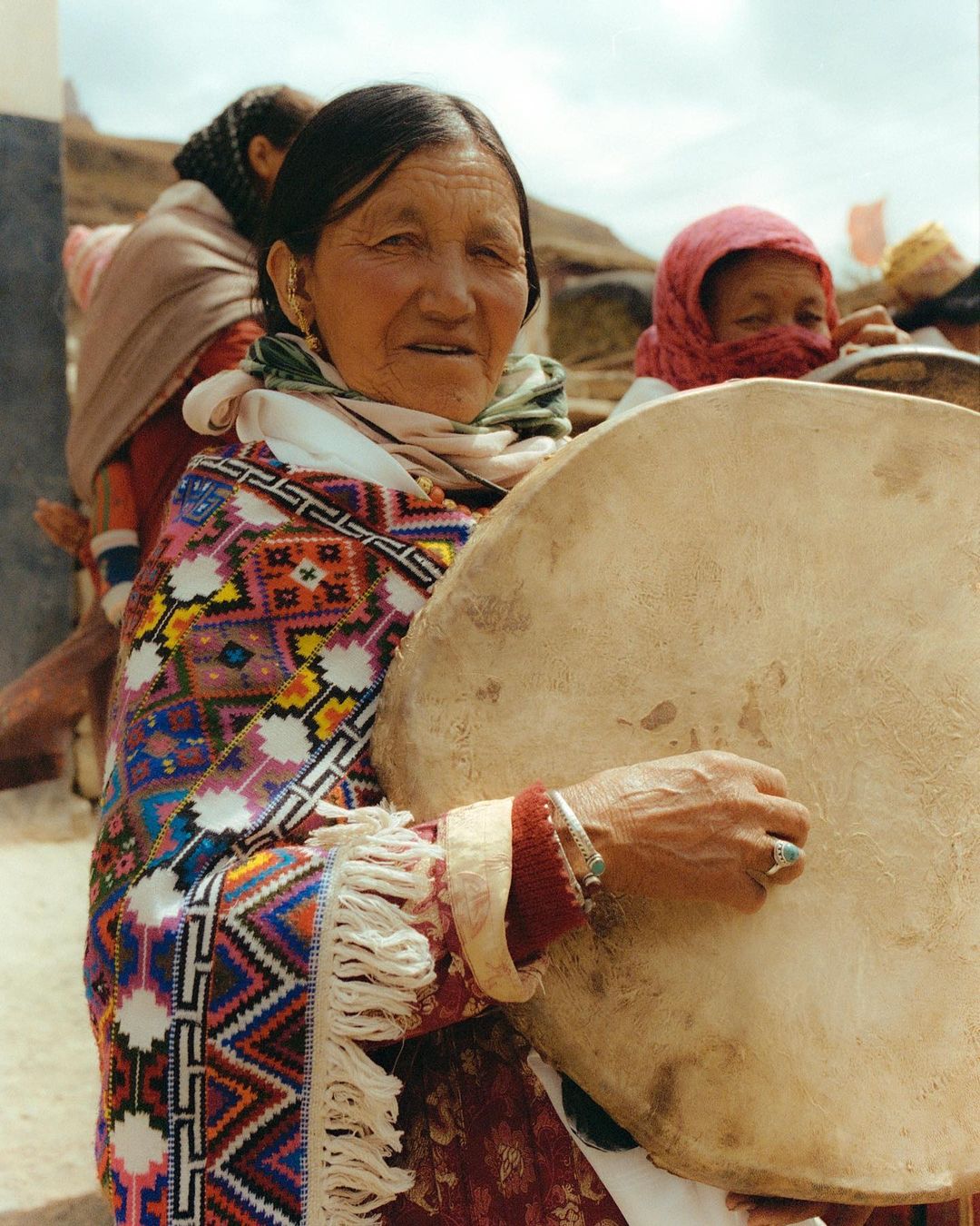 Elderly woman holds a drum outdoors, adorned in a colorful patterned shawl and jewelry, with other individuals blurred in the background.
