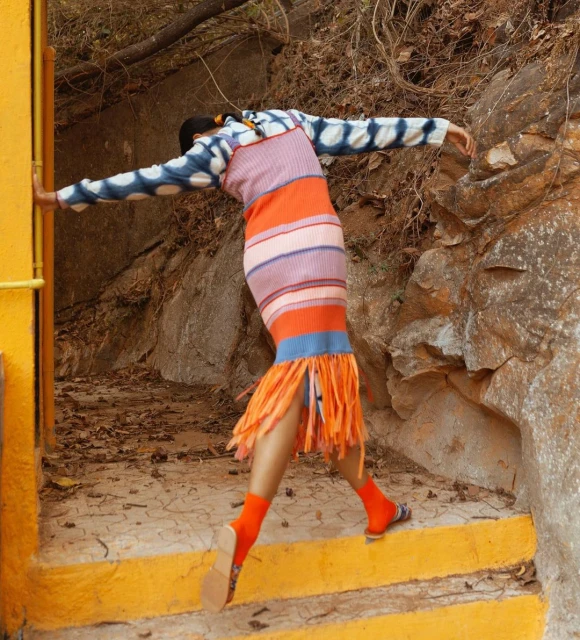 Woman in a colourful dress poses dynamically against a rocky background with orange accents. Bright socks and sandals add to the outfit.