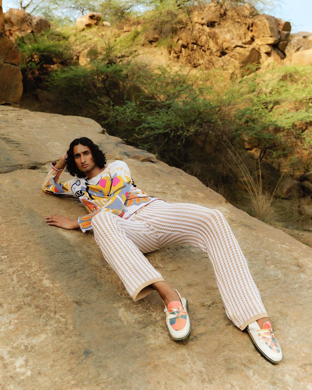 A person lies on a rock in striped pants and a patterned top outdoors. A hillside with scrubby trees is in the background.
