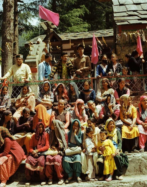 Large group of women, children, and men gathered outdoors near a building and trees with pink flags for an outdoor event.