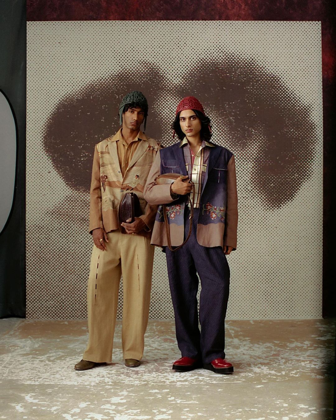 Two stylish young men pose indoors against a backdrop, showcasing modern fashion with patterned jackets, coordinated trousers, and knit caps.